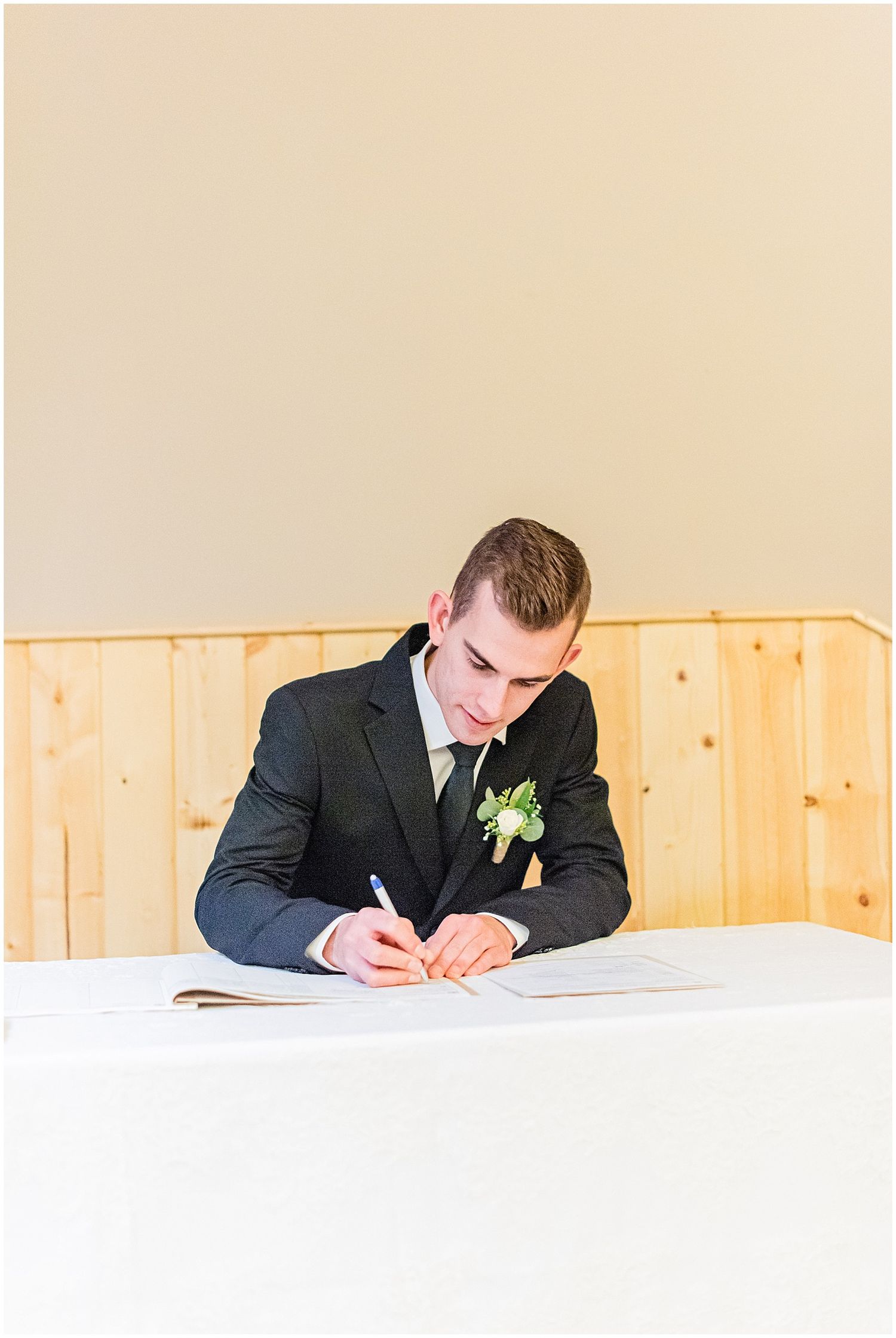Groom in dark suit signing wedding documents at a white draped table with boutonniere.