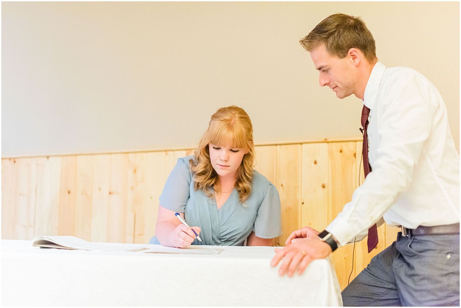 Woman in teal dress signs wedding documents at a table while a man in a white shirt looks on.