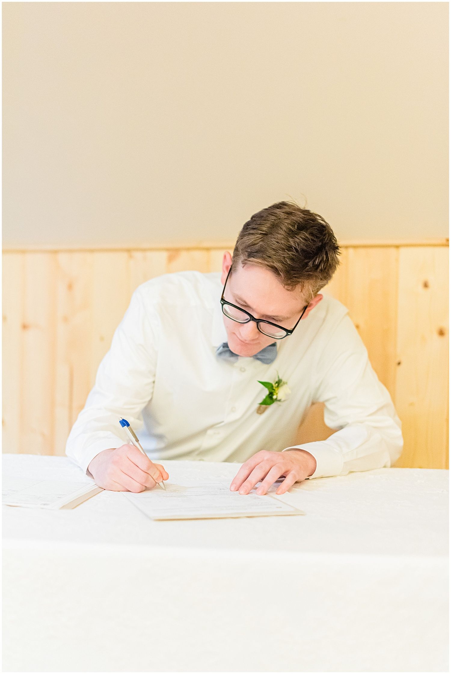 Groom in white shirt and bow tie signs wedding documents at a table.