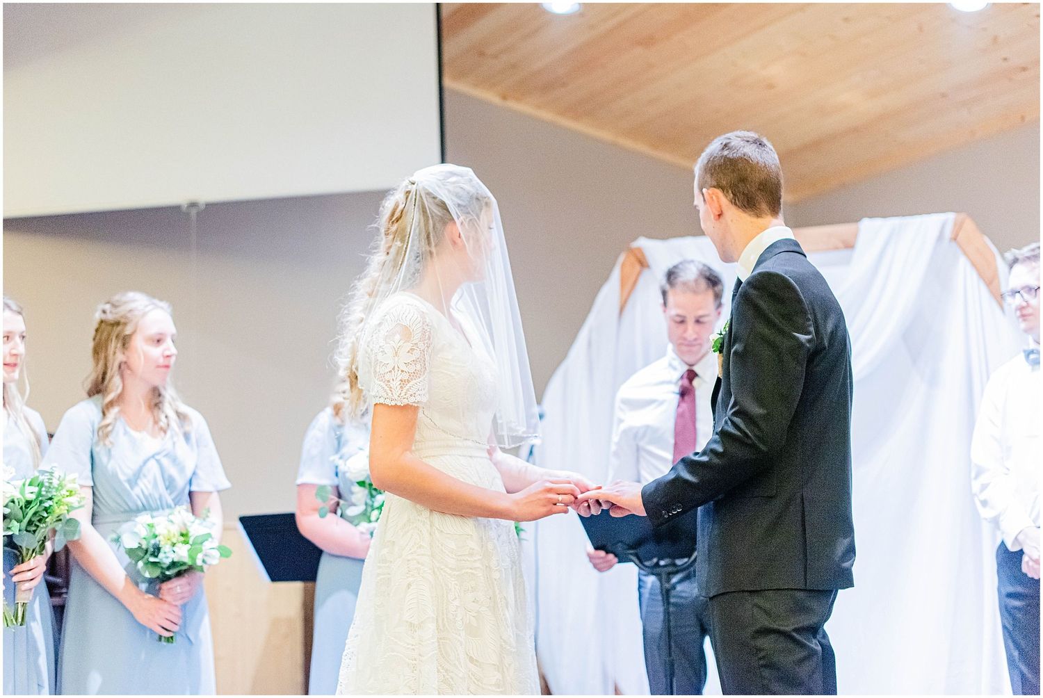 Bride and groom exchanging rings at the altar surrounded by bridesmaids in gray.