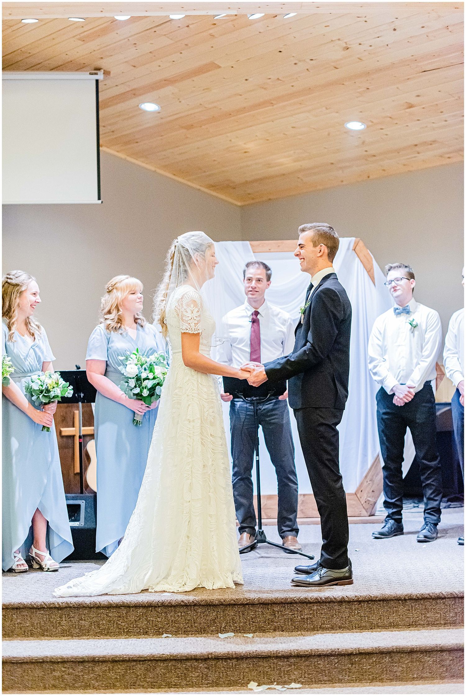 Bride and groom exchange vows on church altar surrounded by bridesmaids in blue dresses and groomsmen.