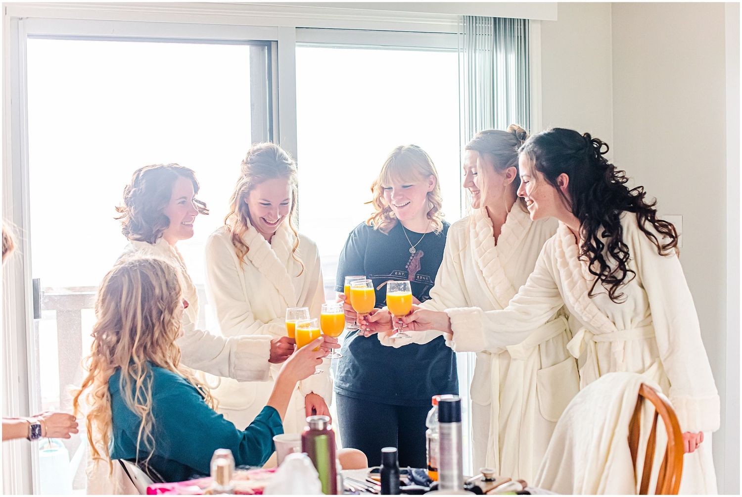 Bridesmaids in white robes toasting with orange juice glasses during bridal party getting ready celebration.
