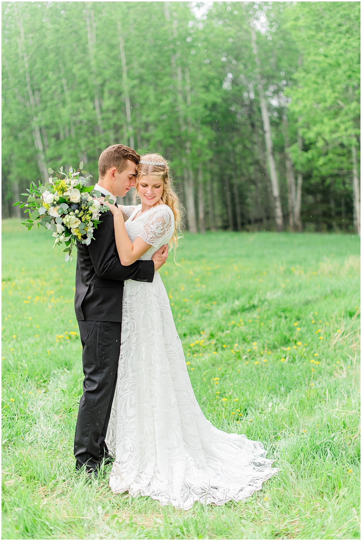Bride and groom embrace in a lush green meadow, her lace gown trailing on the grass with birch trees behind.