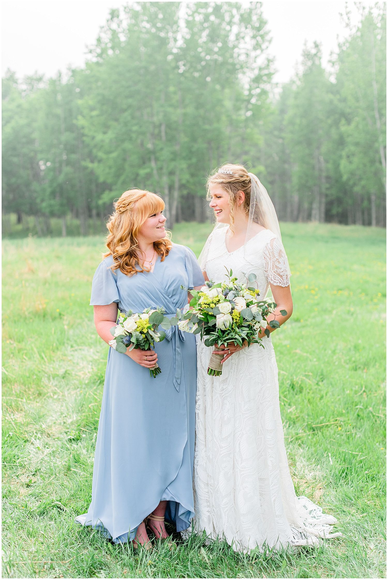 Bride in lace gown and bridesmaid in blue dress holding bouquets outdoors in green field.
