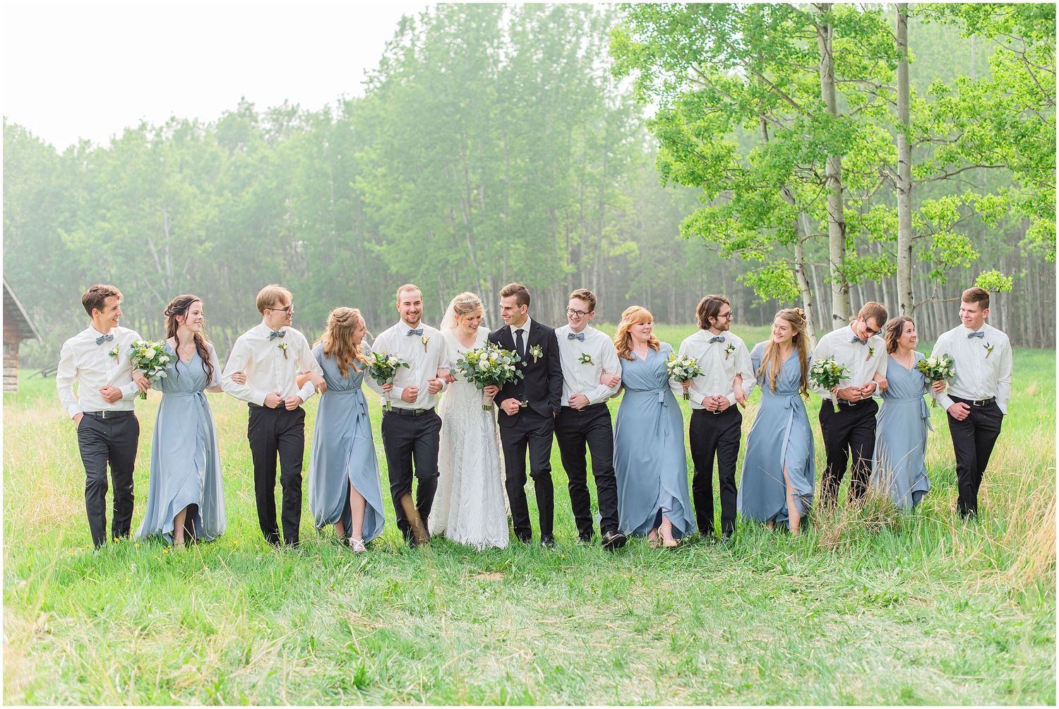 Wedding party in dusty blue dresses and white shirts pose in green field.