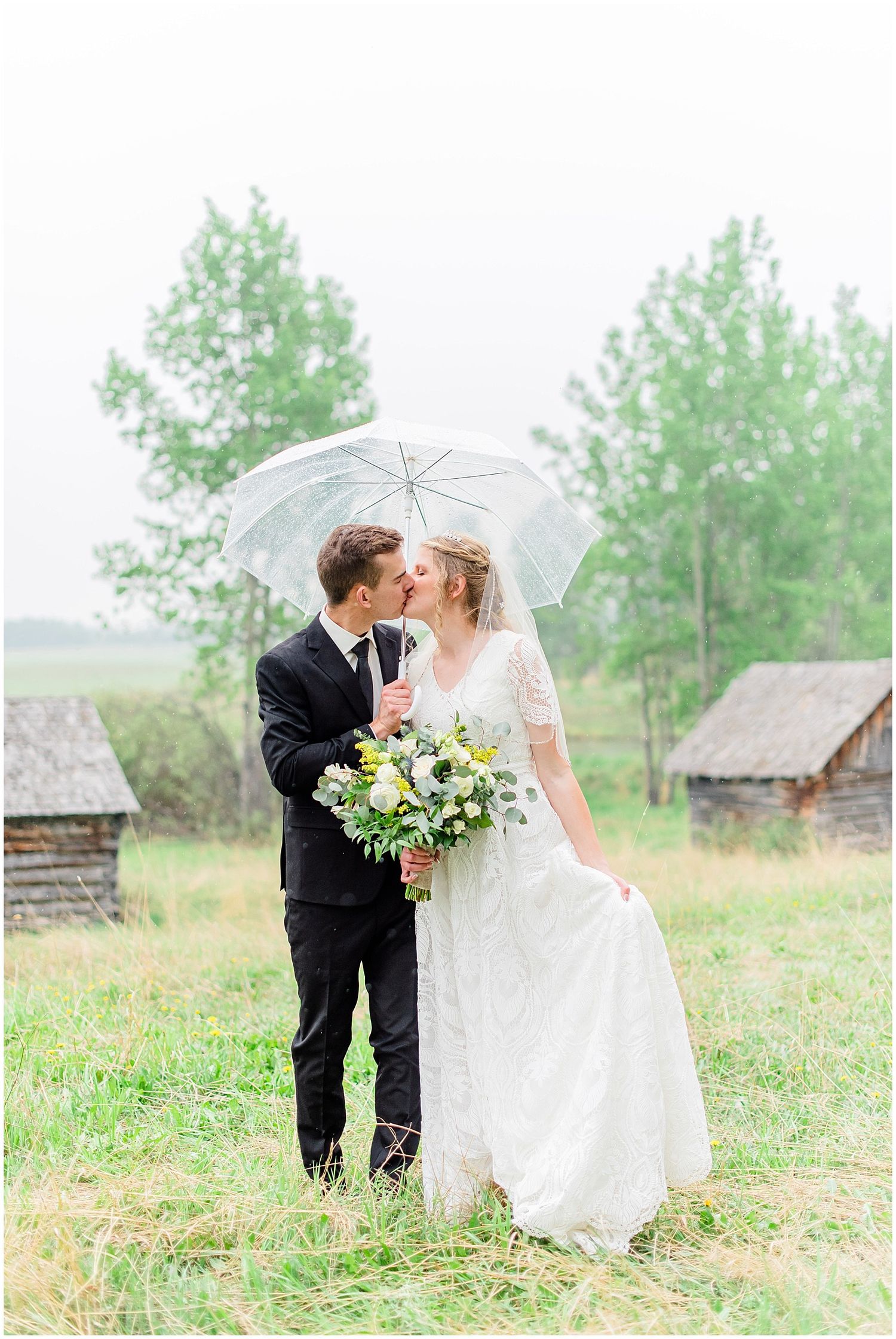 Bride and groom share romantic kiss under clear umbrella in rainy outdoor field setting.