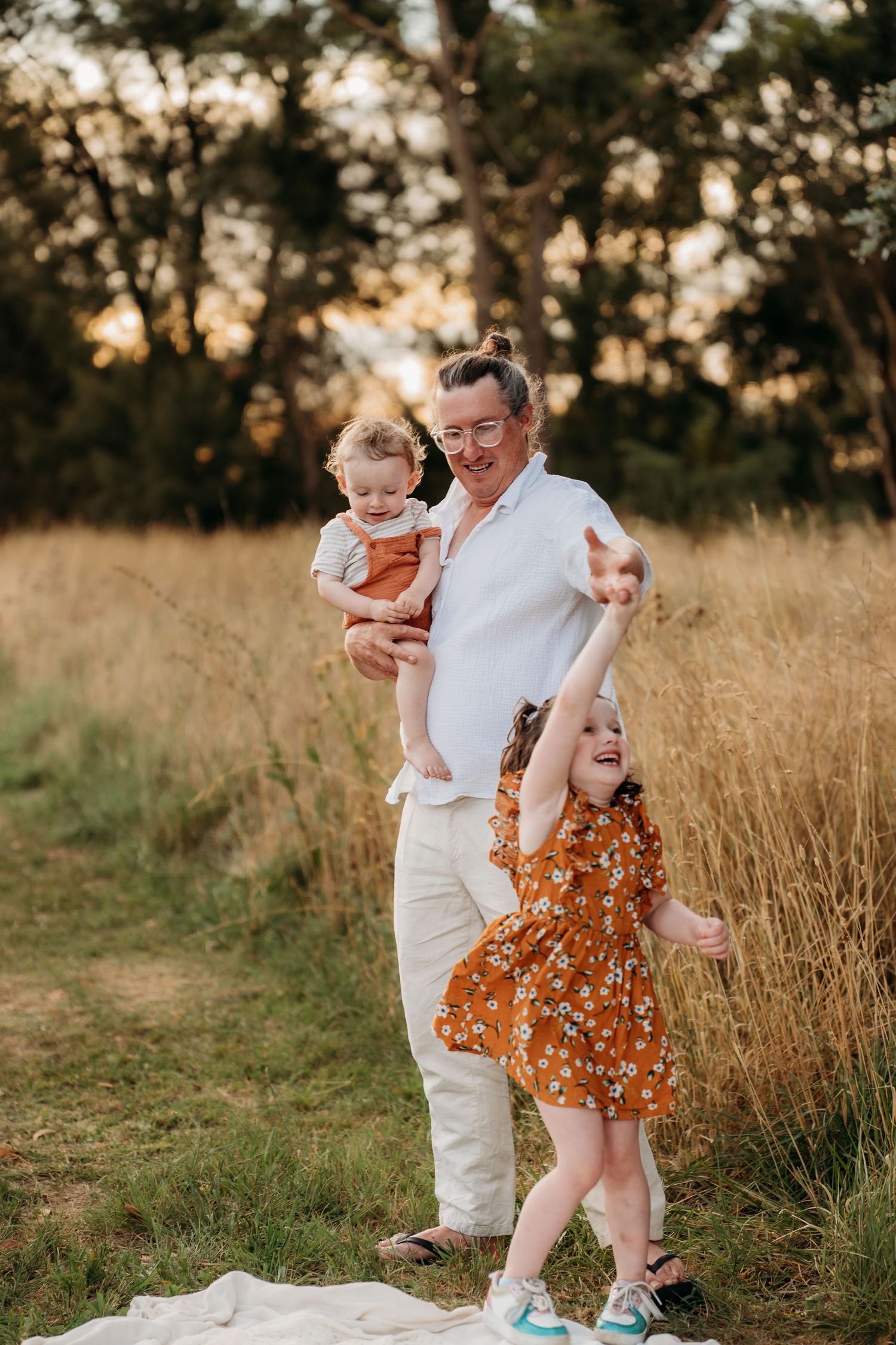 A parent holds a baby while a young girl in a floral dress plays outdoors in a golden field at sunset.