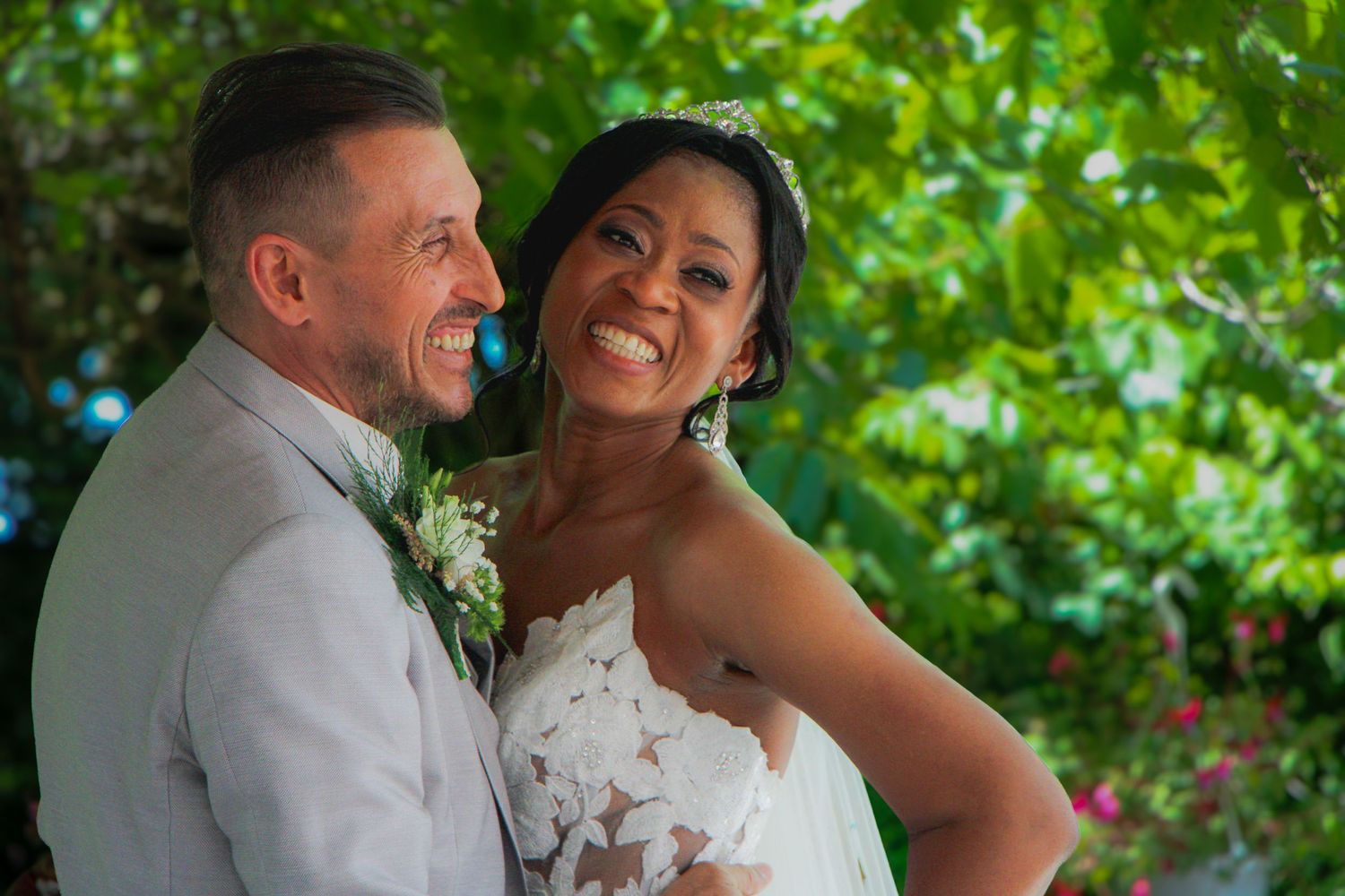 Happy bride and groom smile together outdoors on their wedding day, surrounded by lush green trees.
