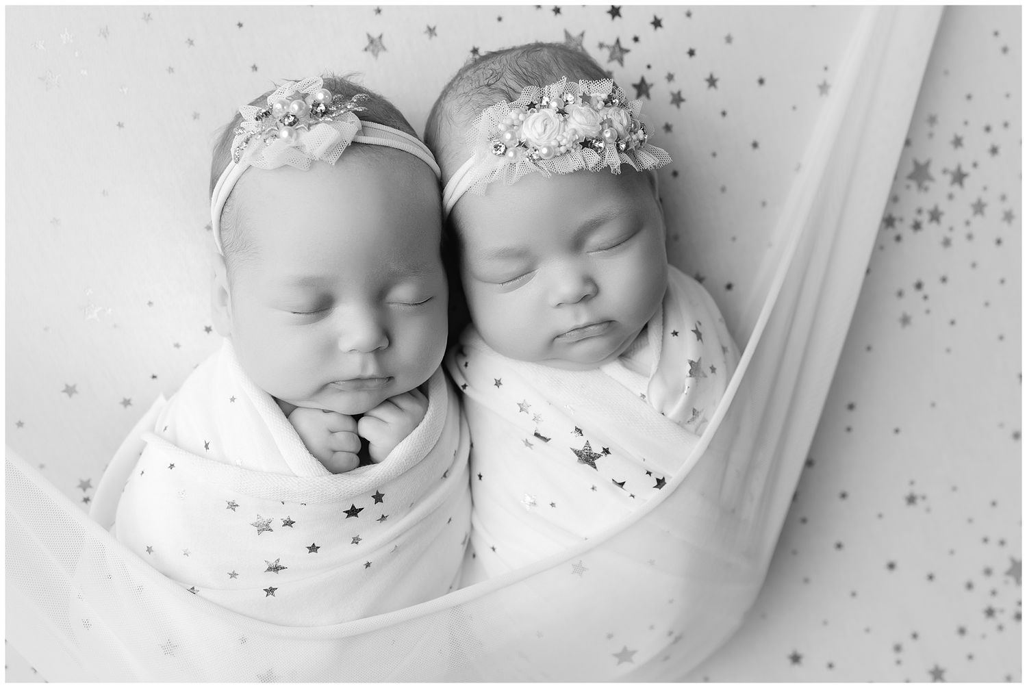 Newborn twin girls, posed swaddled in creamy fabric with stars in a hammock on the same creamy stars fabric background.