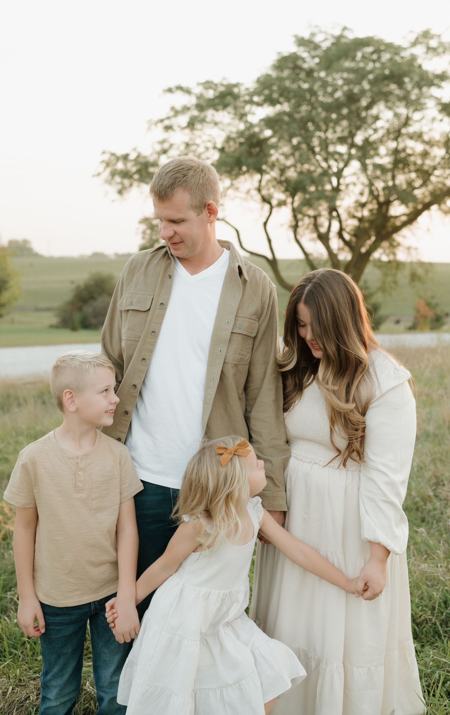 Happy family of four in neutral tones stands together in golden meadow near a peaceful pond.