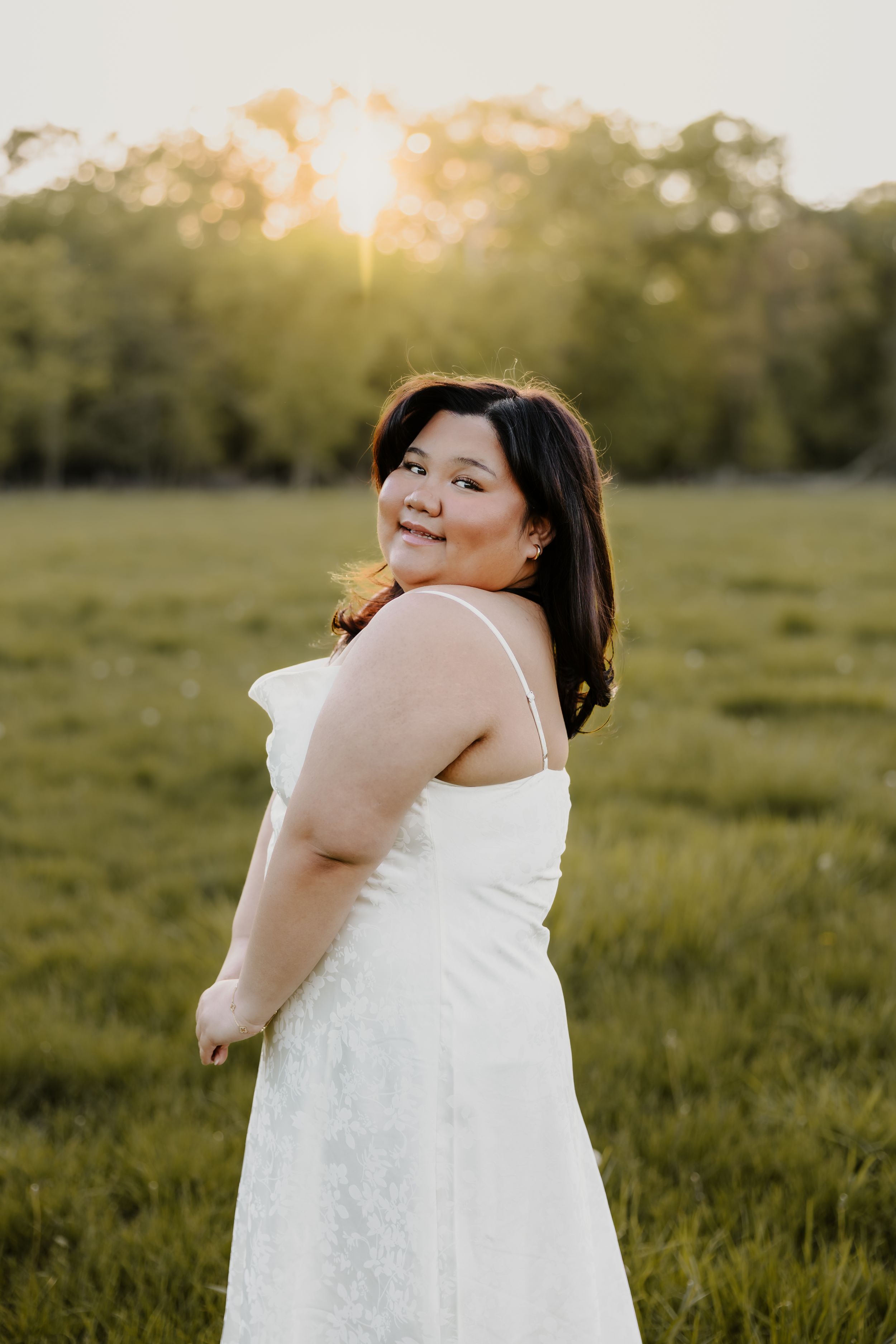 Young woman in white sundress smiling over shoulder in sunlit green field at golden hour.