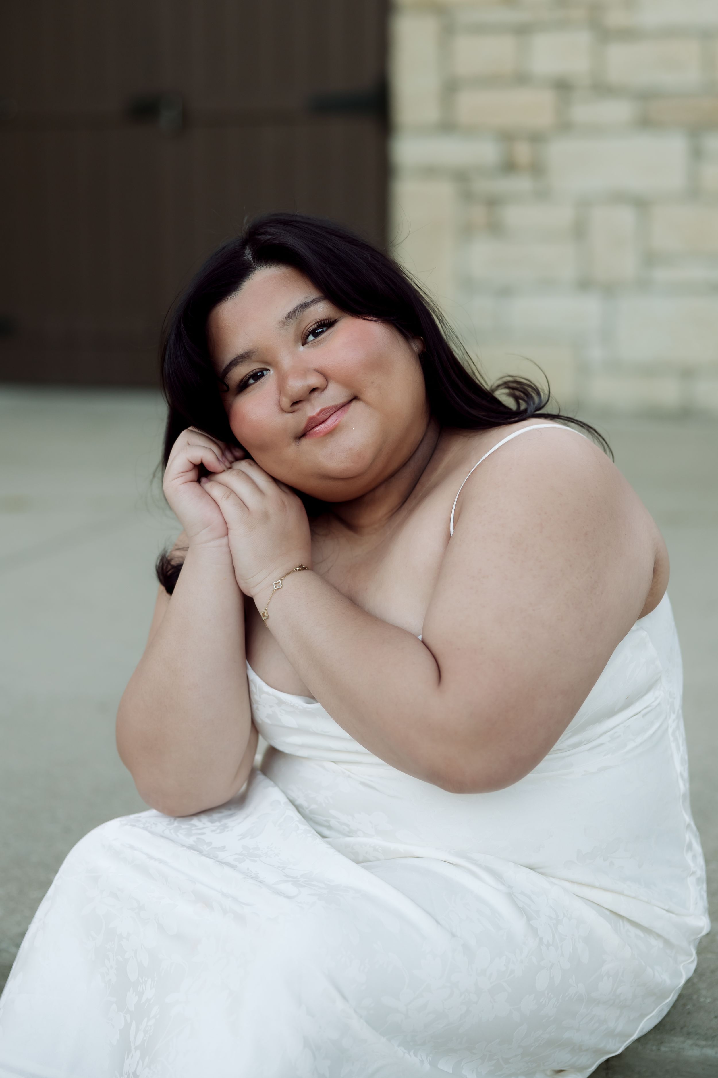 Young woman in white dress sitting and smiling with hands clasped near face against stone wall.