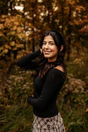 Young woman in black off-shoulder top and plaid skirt smiling outdoors among autumn foliage.
