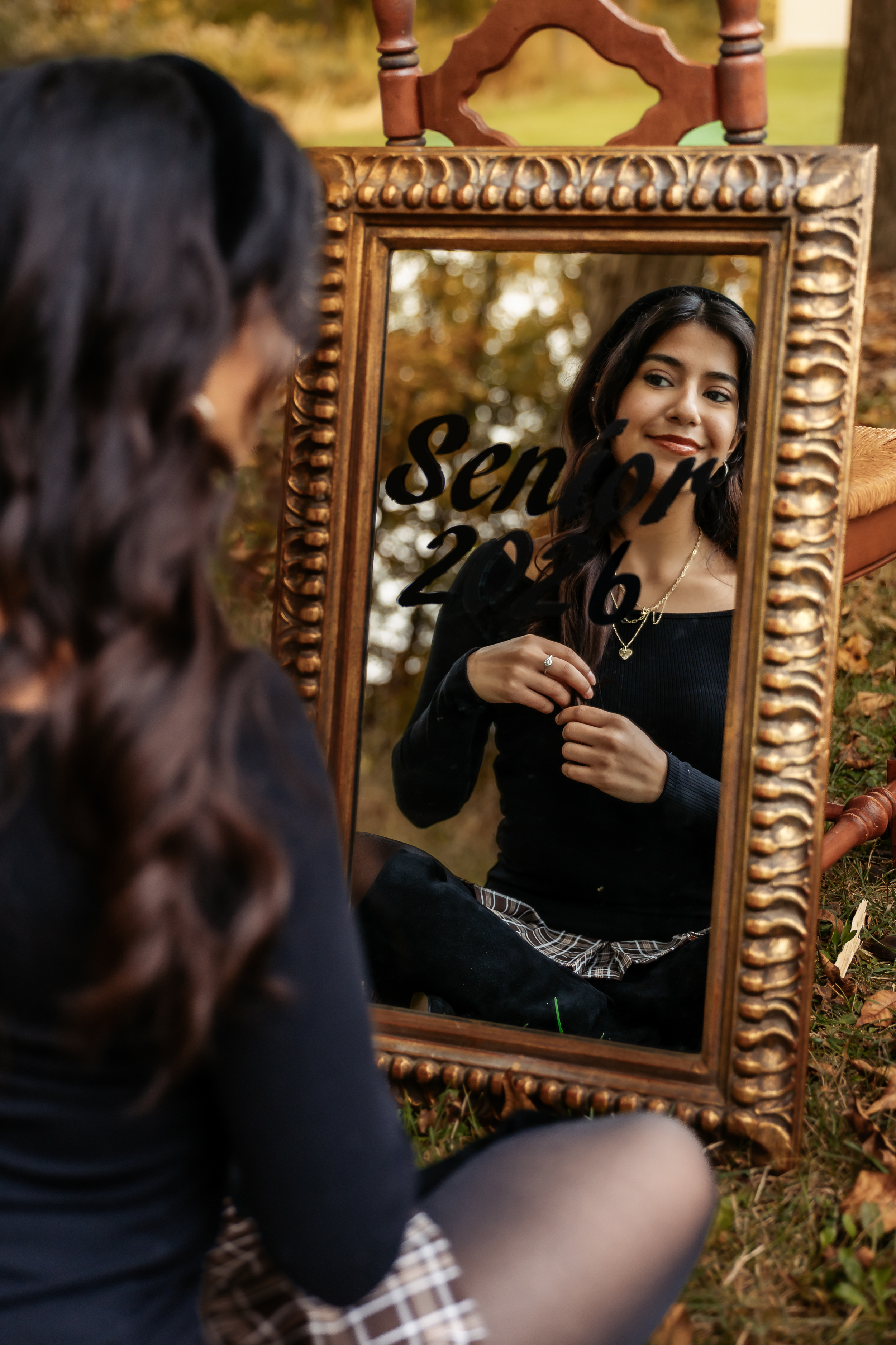 A young woman sits before an ornate gold mirror reflecting her smile, with "Senior 2026" text overlay, outdoors in fall.