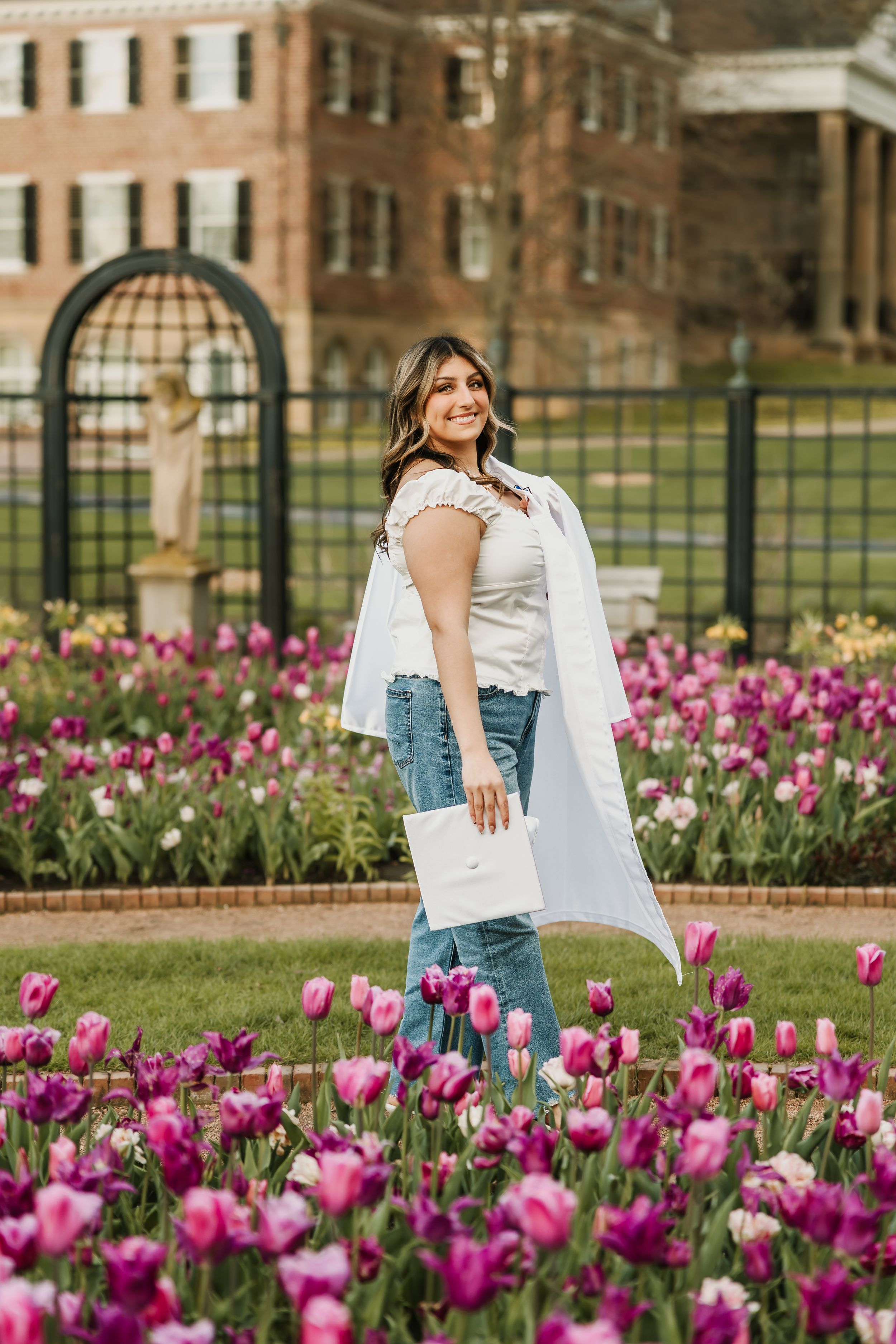 Young woman in white graduation gown holding diploma among pink tulips in garden with brick building backdrop.