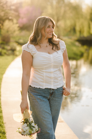 Woman in white top and jeans holding flowers walks by a sunlit pond in a lush green park.