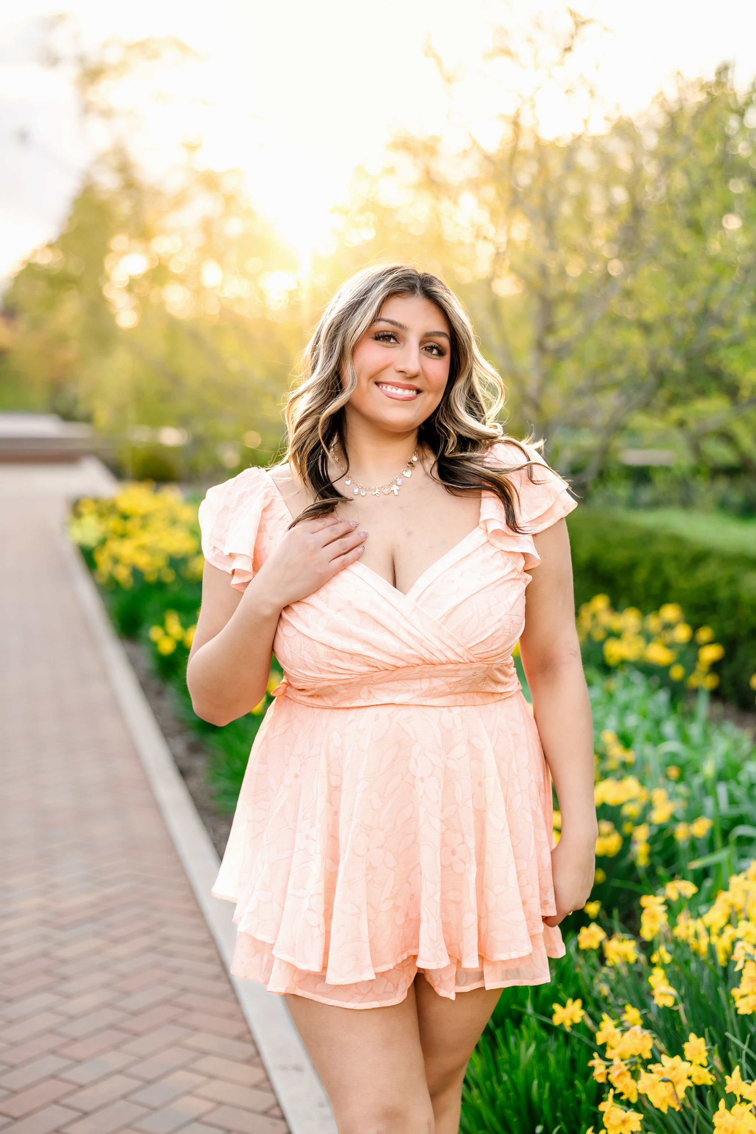 Young woman in peach romper smiling outdoors among yellow daffodils during golden hour in a garden pathway.