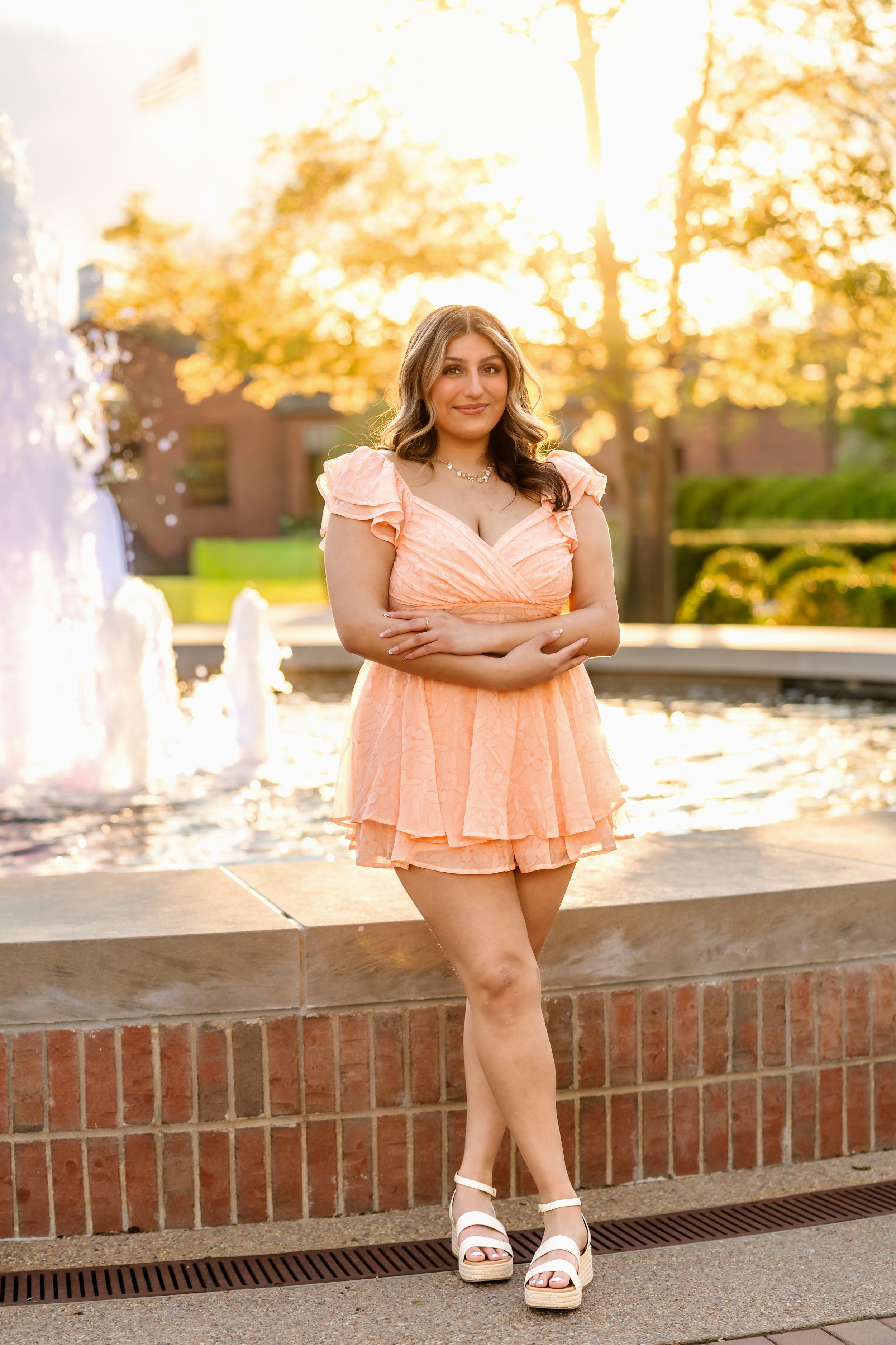 Young woman in peach ruffle mini dress poses by fountain at golden hour in outdoor setting.