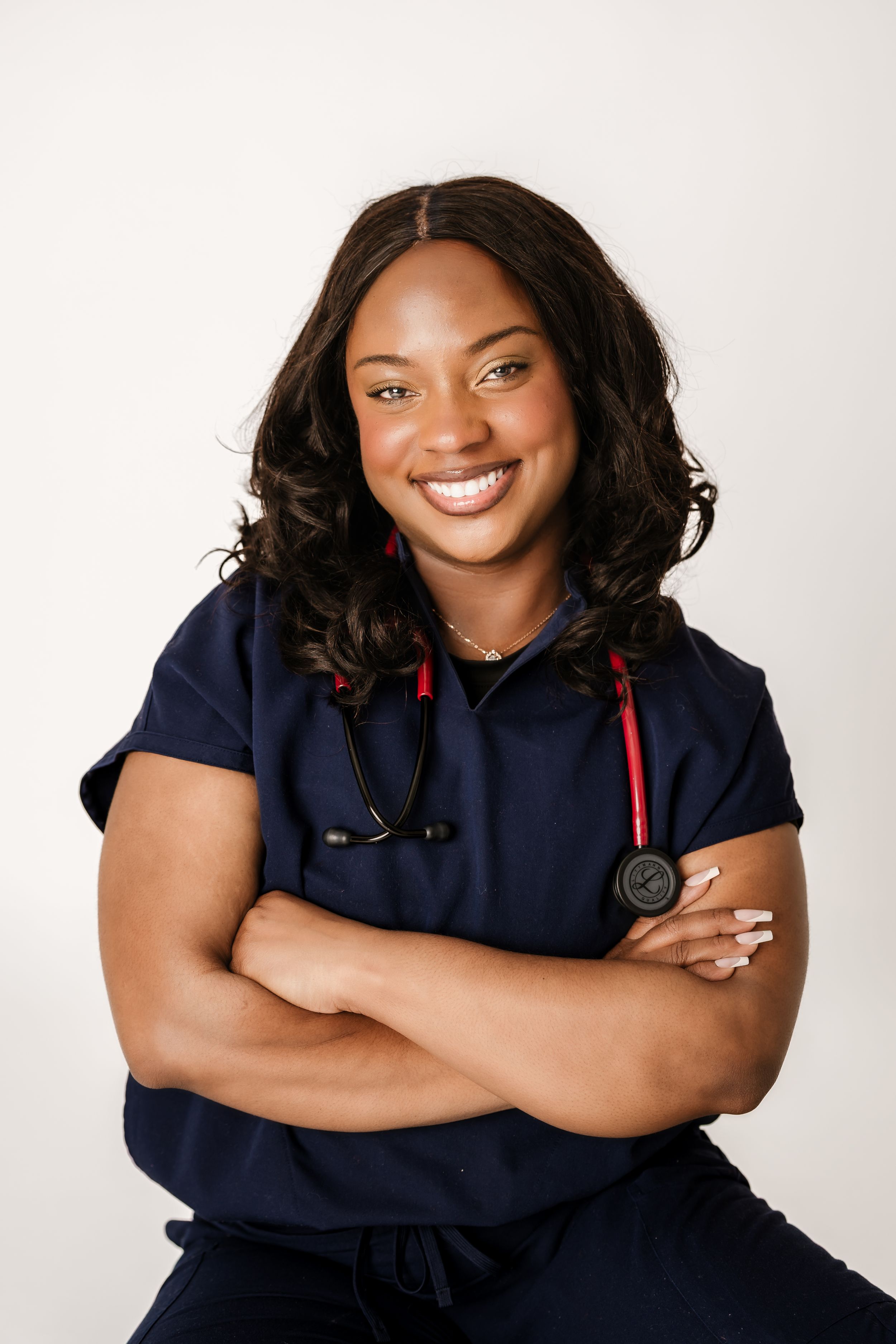 Smiling female nurse in navy blue scrubs with red stethoscope, arms crossed, posing confidently against white background.