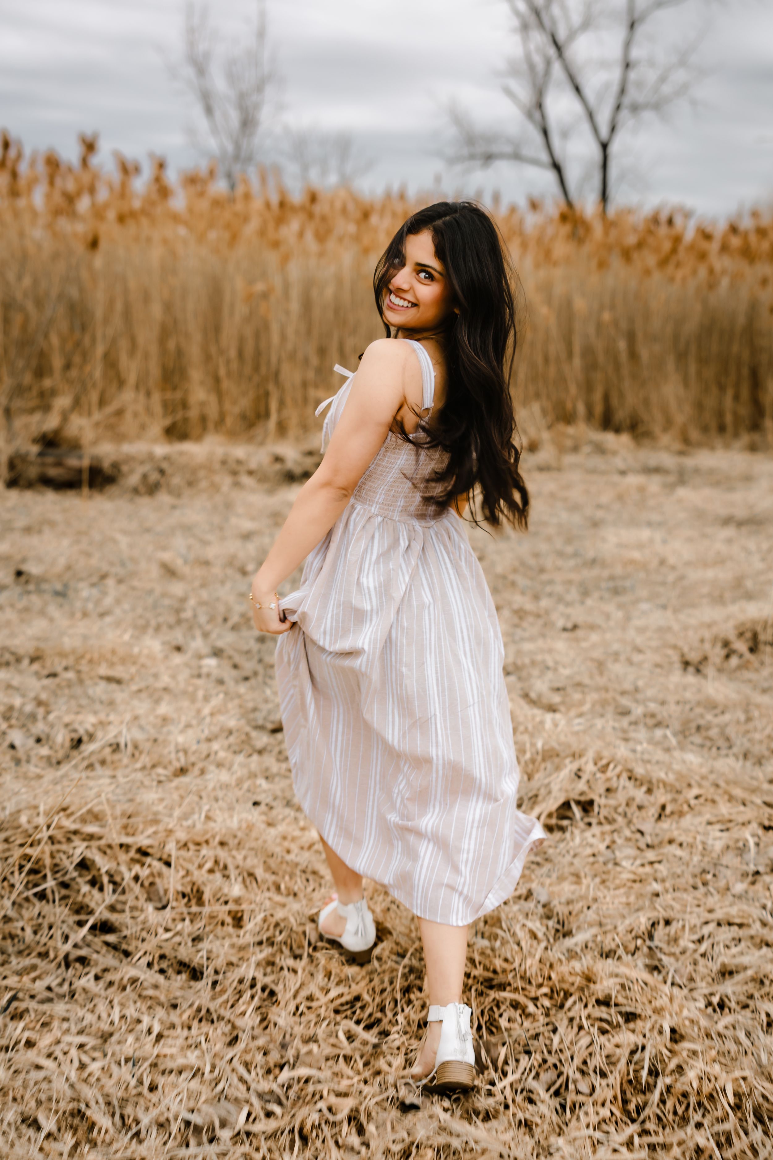 A young woman in a striped midi dress smiles over her shoulder in a golden autumn field.