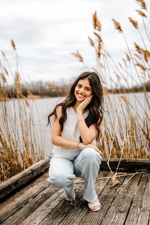 Young woman smiling on wooden dock by lake, surrounded by tall golden reeds, wearing light gray top and blue pants.