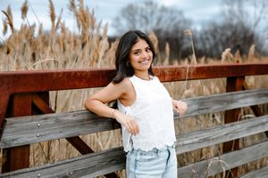 Young woman in white lace top and jeans smiling outdoors by rustic fence with tall golden grasses.