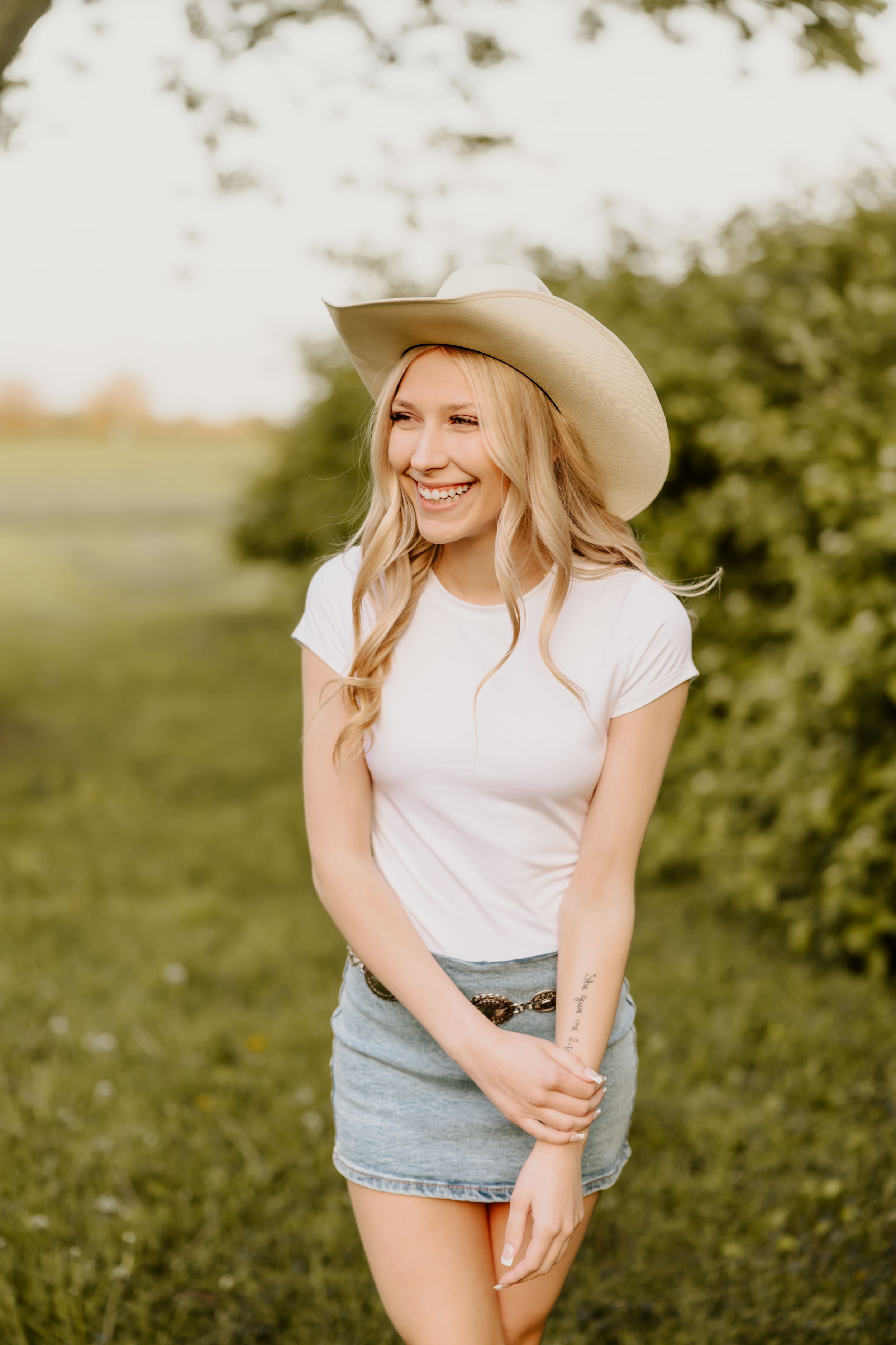 Smiling blonde woman in white tee, denim skirt, and cowboy hat poses outdoors in a sunny green field.
