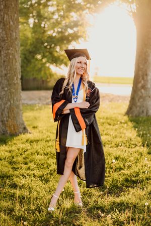 Smiling blonde graduate in black cap and gown with orange tassel poses outdoors in golden sunlight among trees.