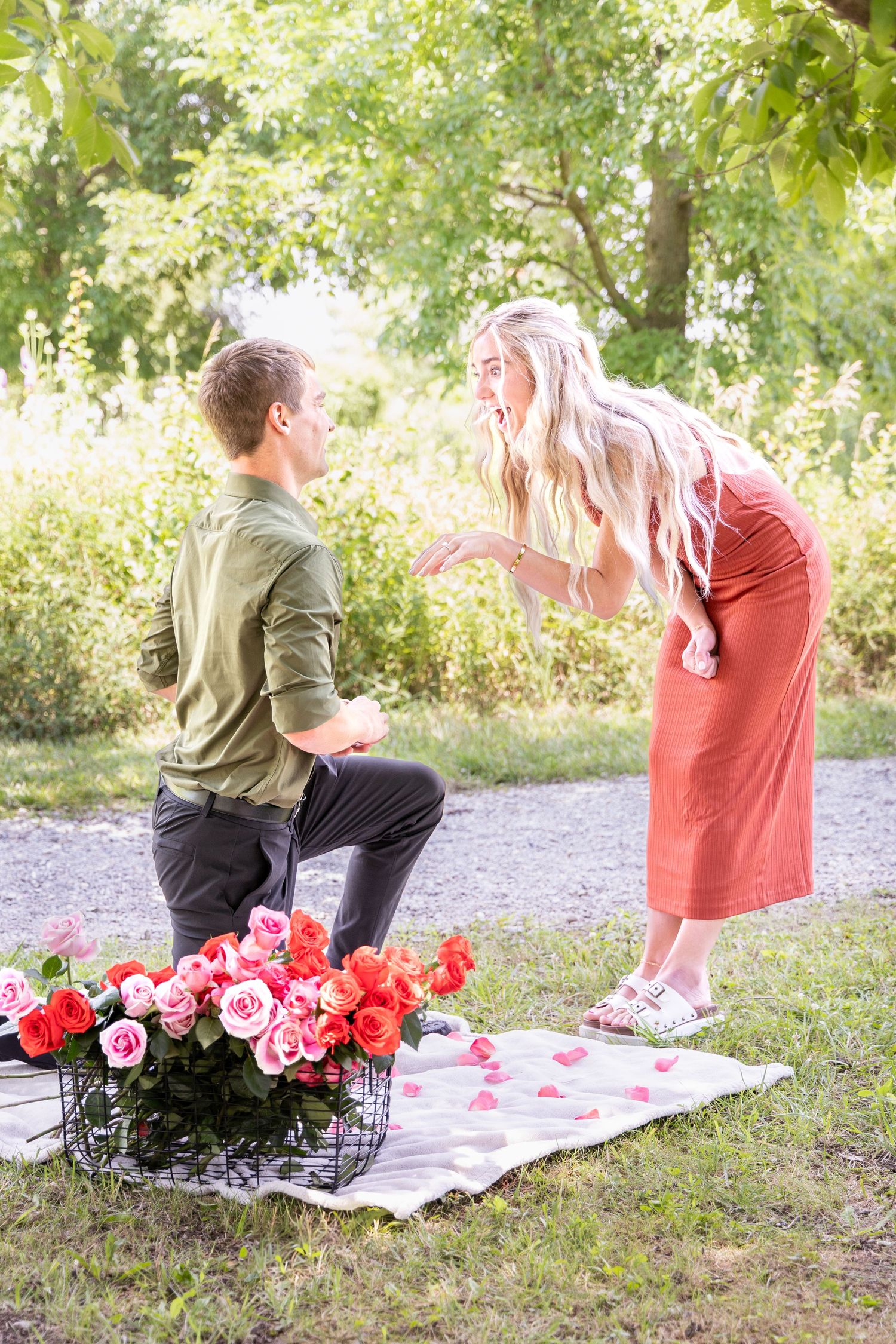 Man on one knee proposing to blonde woman with roses on blanket at Voice of America Park