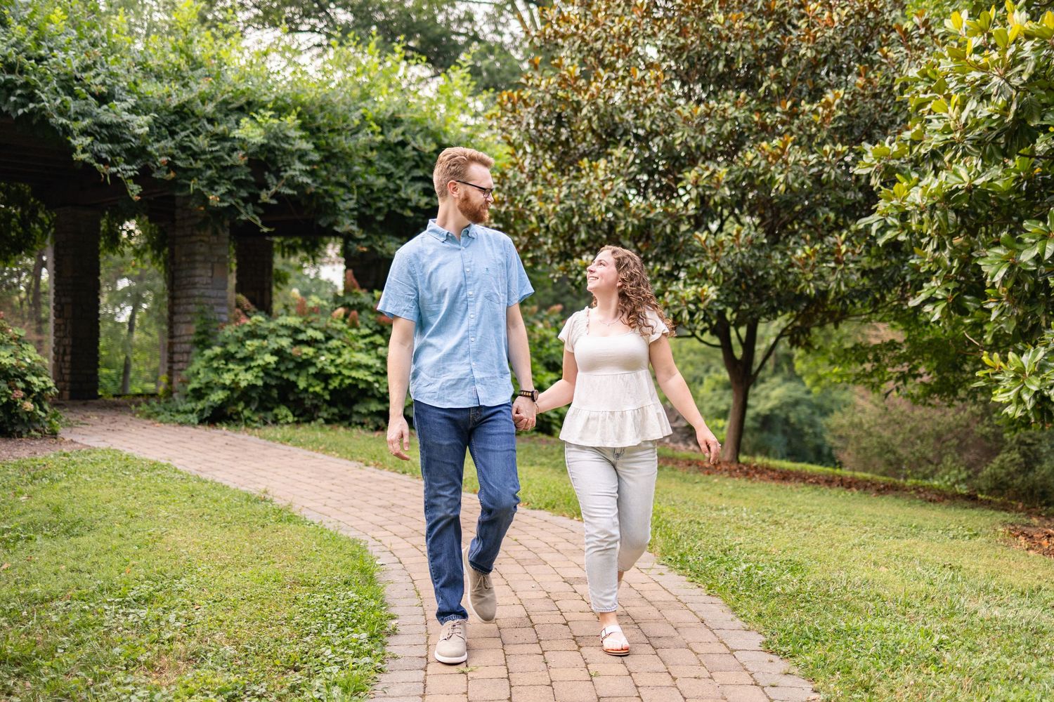 A couple holds hands and walks along a brick garden path surrounded by lush green trees and shrubs.