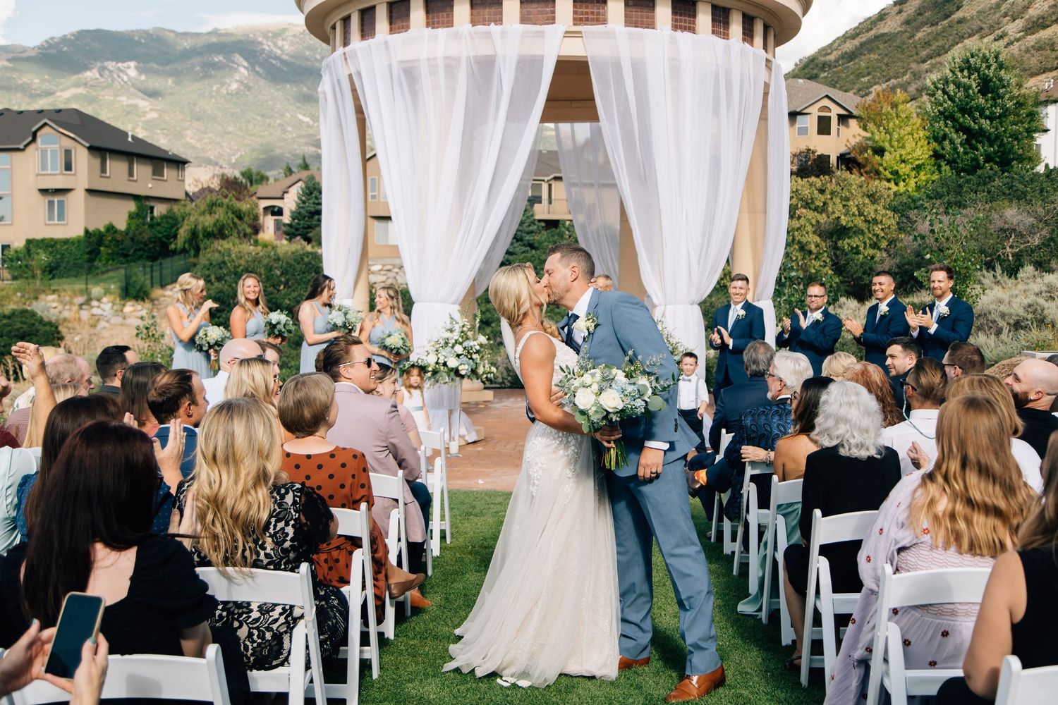 Couple sharing a kiss at outdoor wedding ceremony under draped gazebo with mountain backdrop and guests seated.
