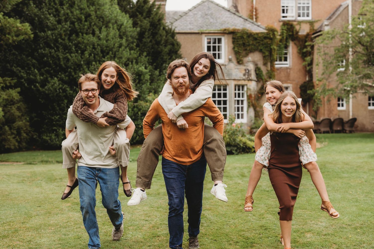Three couples laugh outdoors, with women riding piggyback on men, in front of a brick manor house on green lawn.