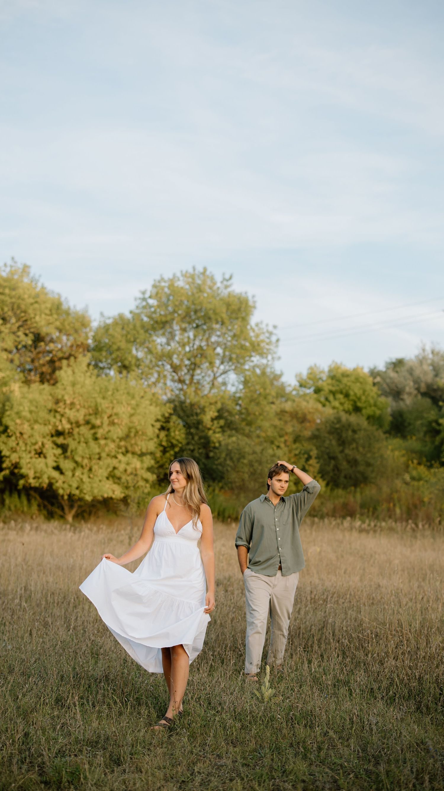 Couple walking in a golden field, woman in white dress, man in green shirt, surrounded by lush trees under a blue sky.