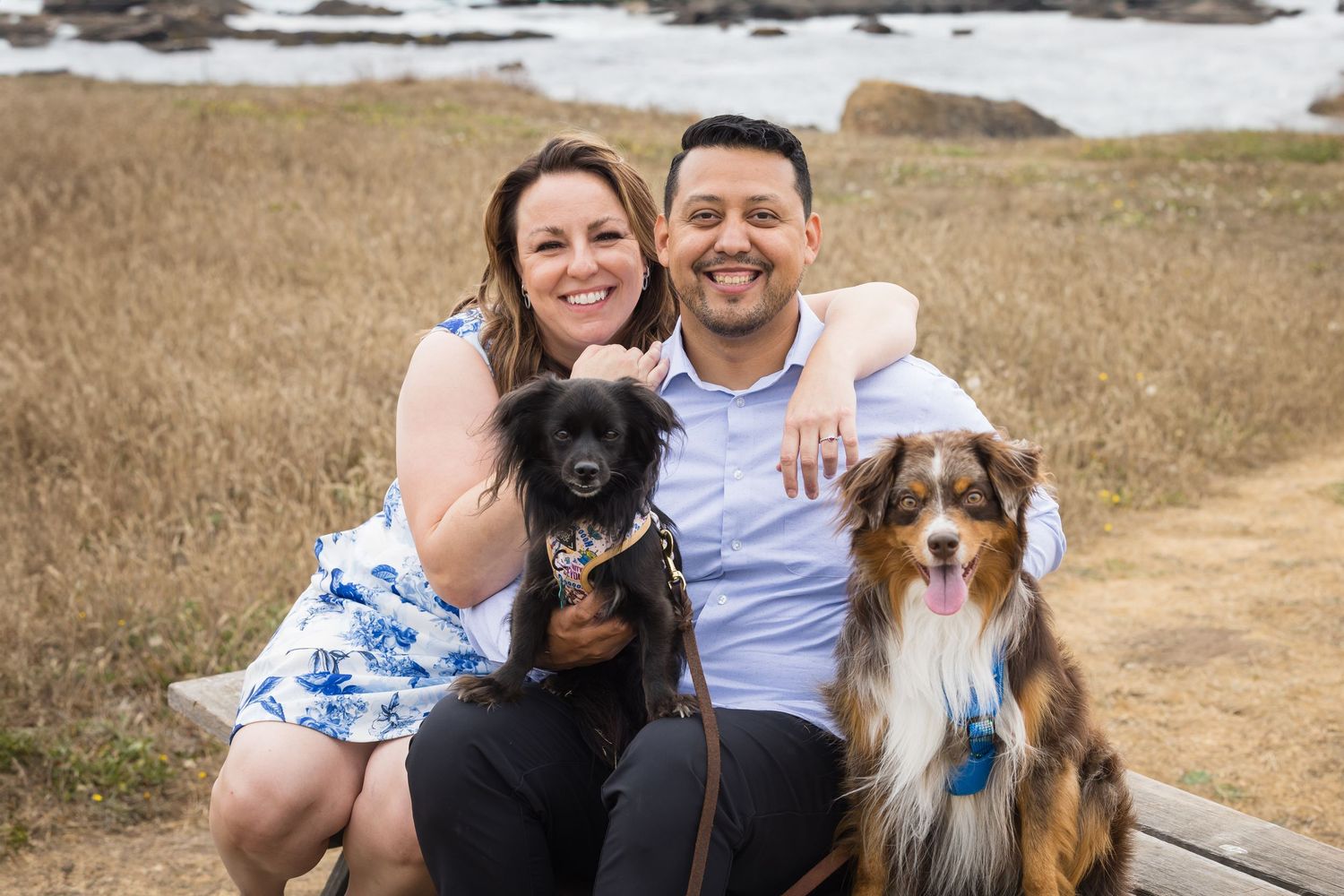 Smiling couple sitting outdoors with a small black dog and an Australian Shepherd near a coastal field.