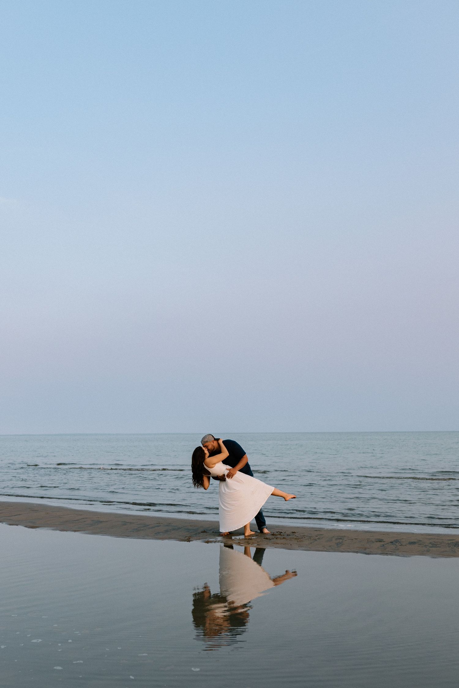 Couple dancing romantically on a beach at dusk, reflected in wet sand with calm ocean in the background.