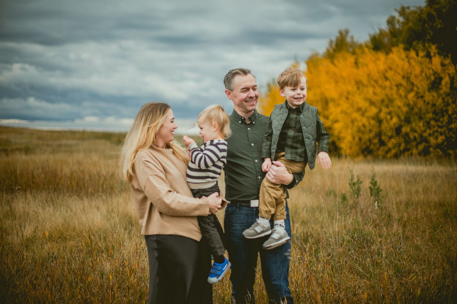 Family of four posing in golden autumn field with dramatic cloudy sky and yellow trees in background.