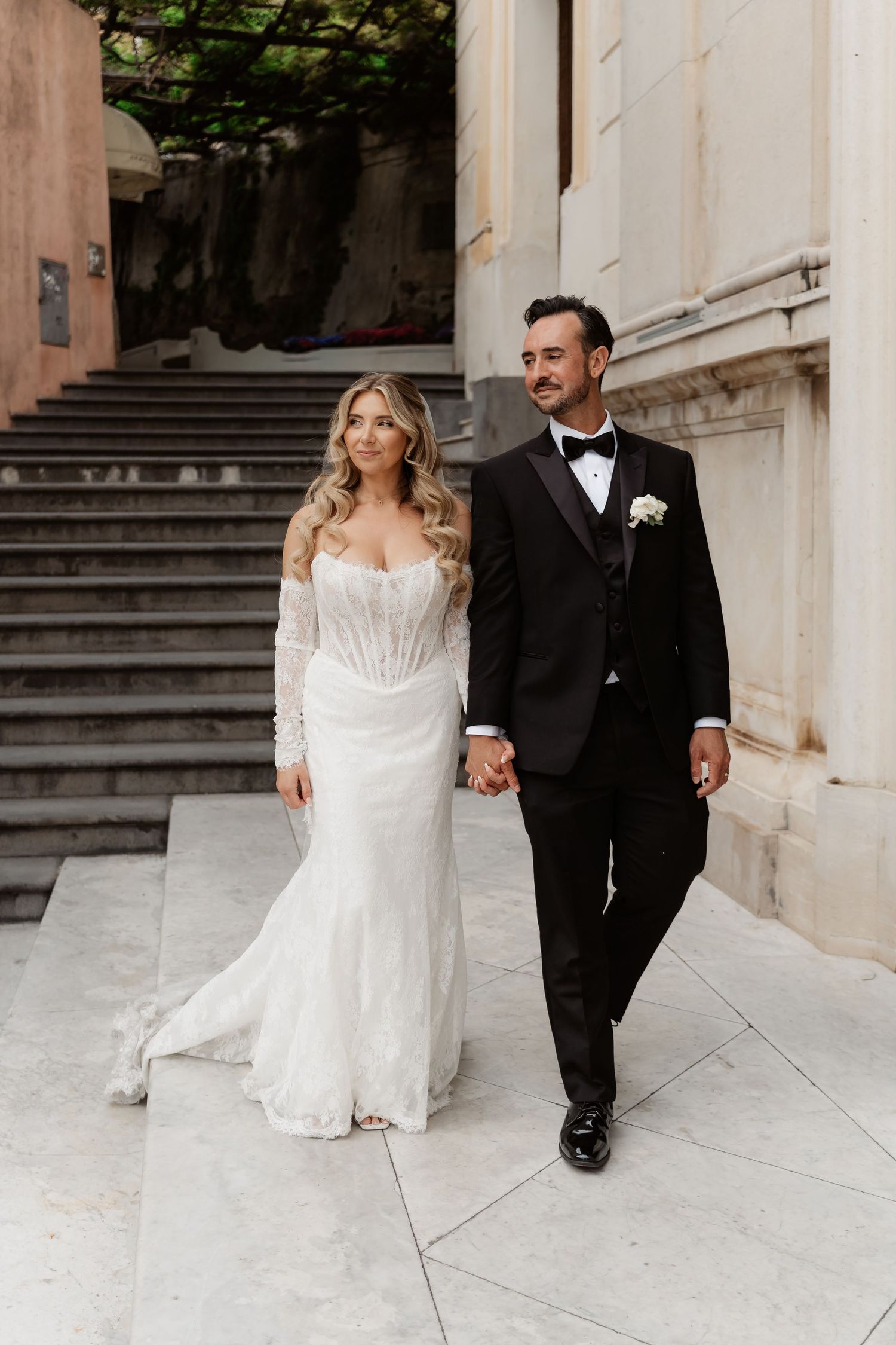 Bride in white lace gown and groom in black tuxedo walk hand in hand at elegant wedding venue.