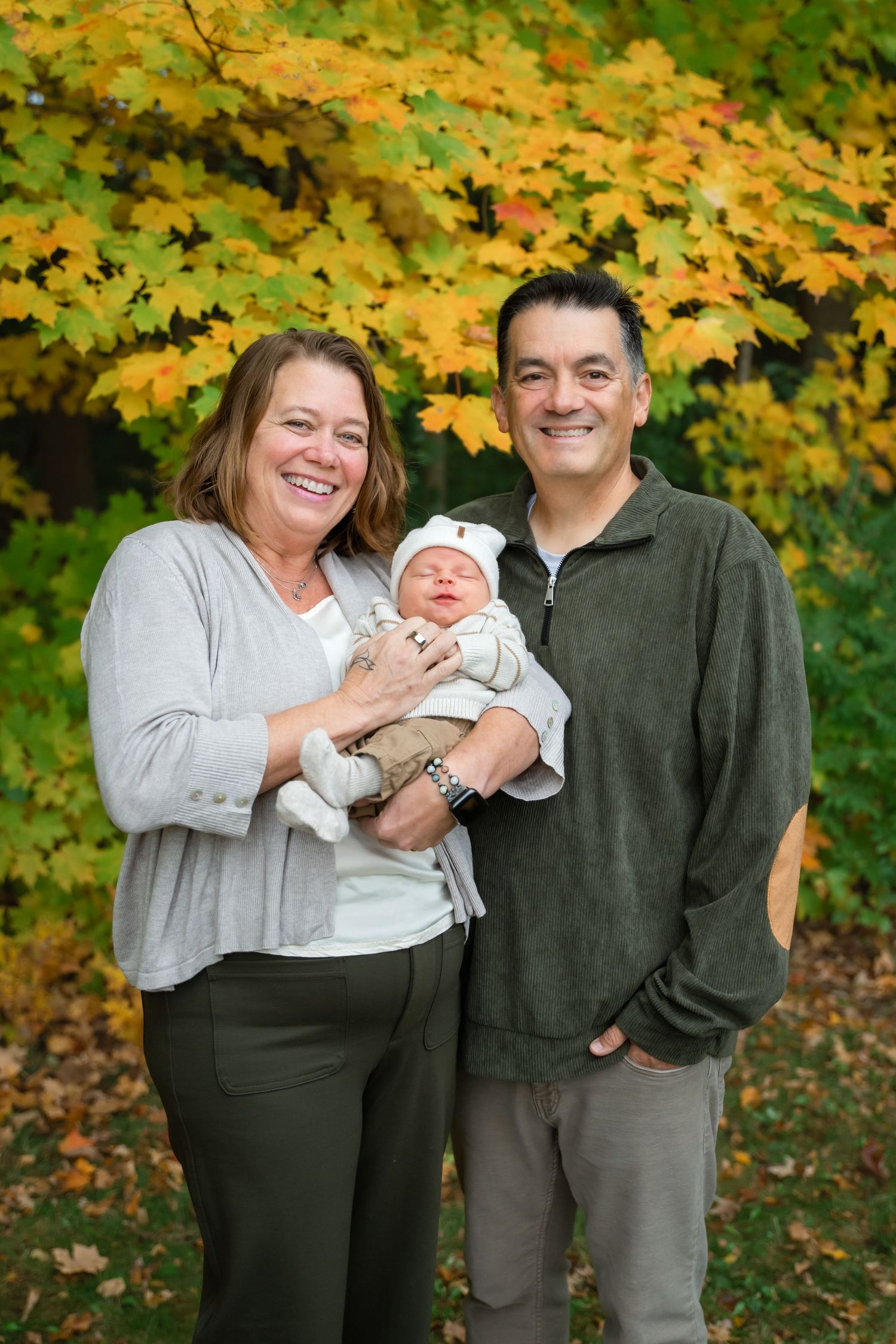 Smiling couple holding a newborn baby outdoors with vibrant autumn foliage in the background.