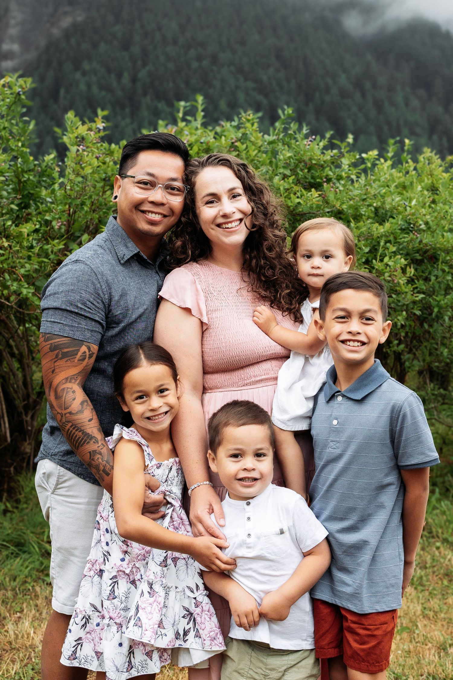 A happy mixed-race family of six poses outdoors with lush green trees and misty mountains in the background.