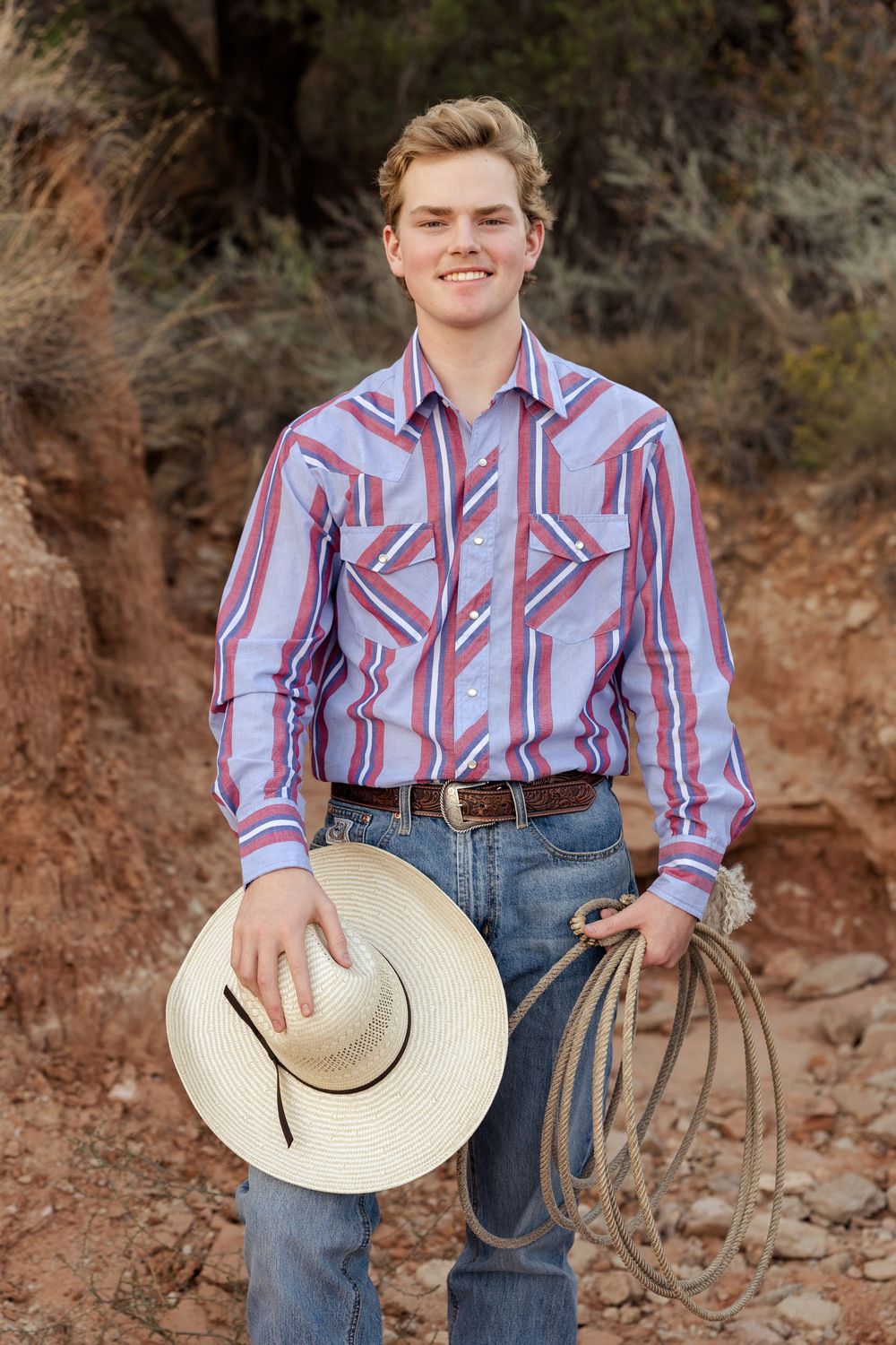High school boy smiles at the camera holding his hat and rope for his senior portraits in Palo Duro Canyon during golden hour.