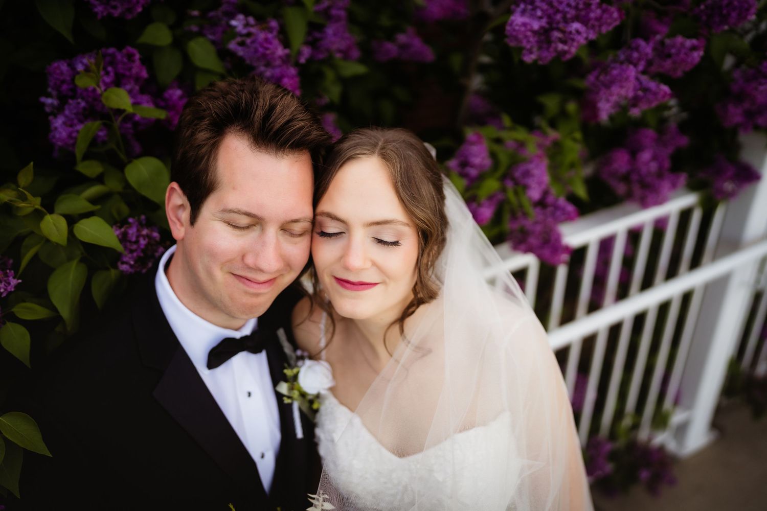 Bride and groom embracing tenderly in front of blooming purple lilac bushes, bride wearing veil and lace gown.