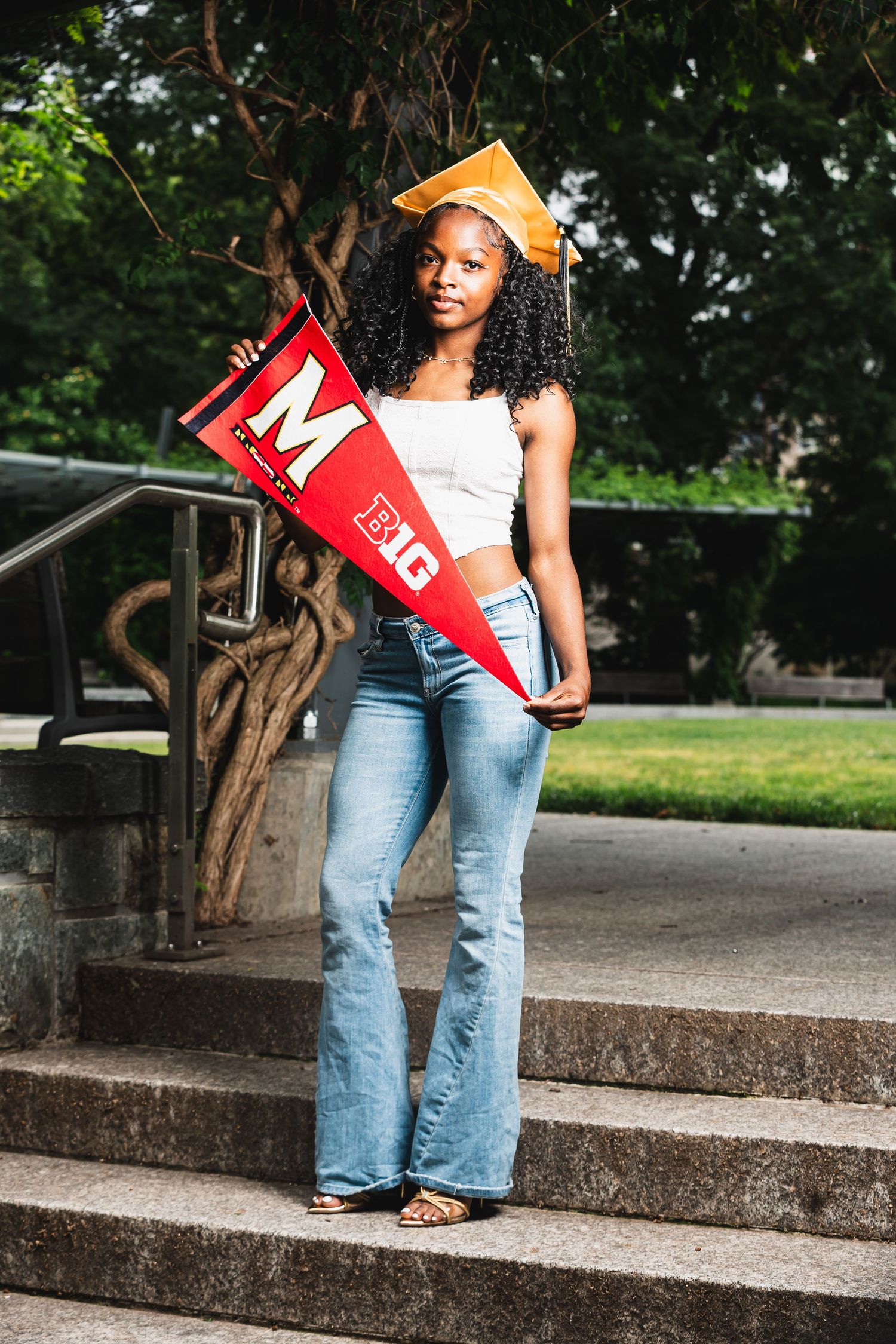 A young woman in jeans and graduation cap holds a University of Maryland pennant flag on outdoor steps.