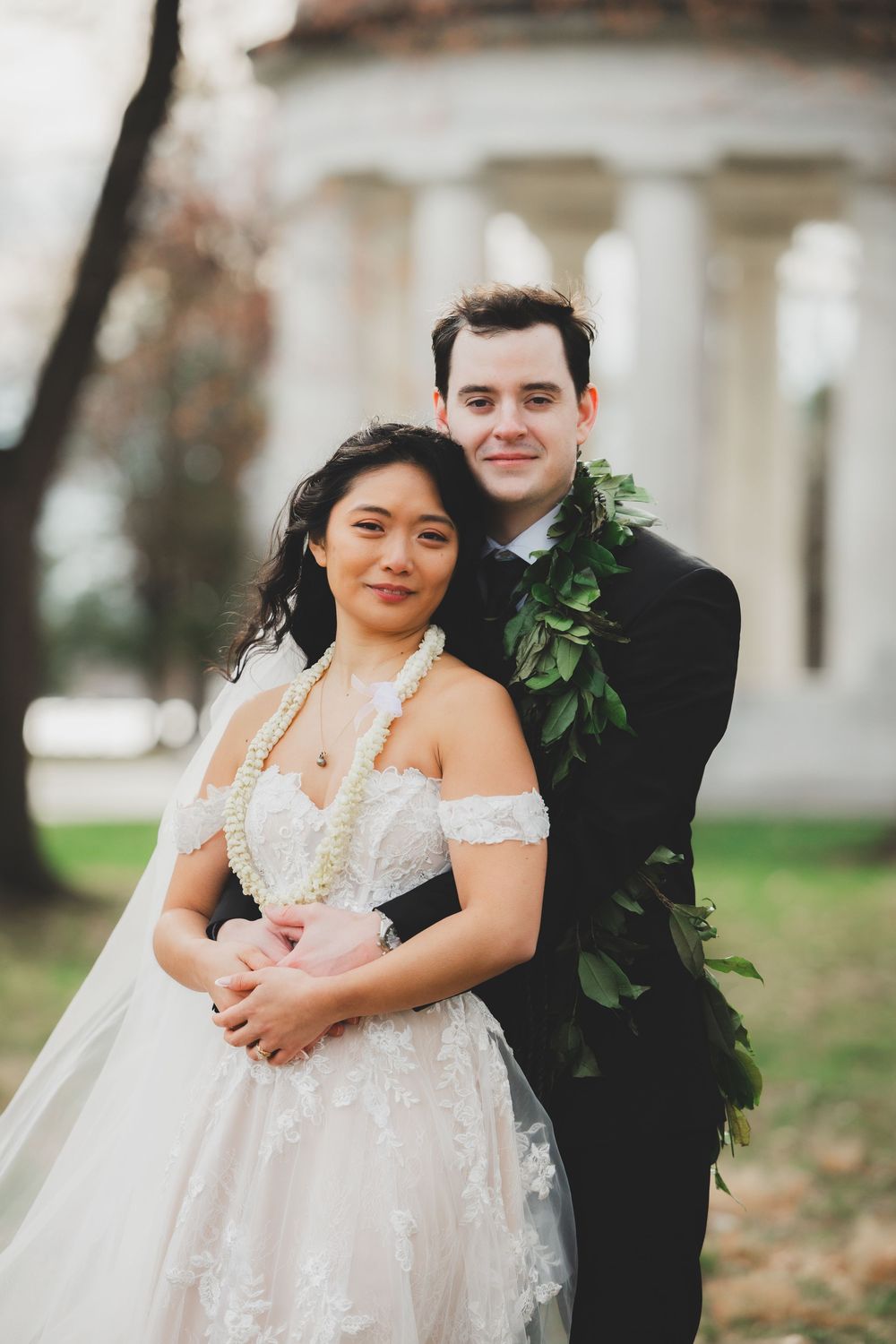 Bride and groom posing outdoors in wedding attire, she in lace gown, he in black suit with green lei.