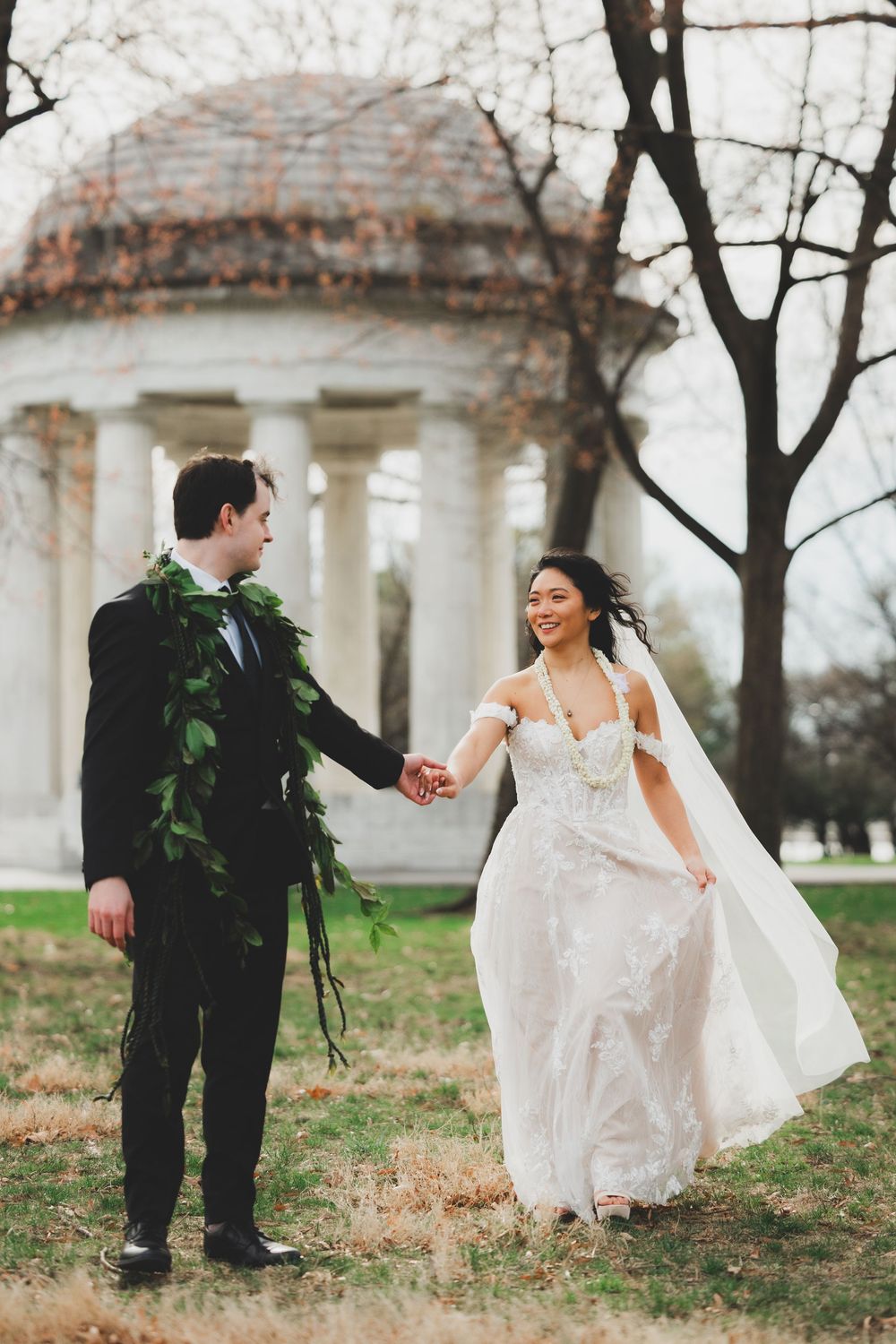 A bride and groom hold hands in front of the dc war memorial, she in lace gown, he in black suit with greenery.