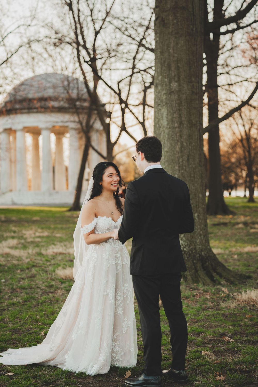 Bride in floral gown and groom in black tux share a moment near a tree, with a domed monument in the background.