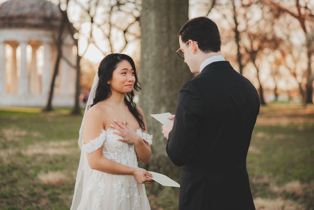 Bride and groom exchange wedding vows outdoors in a park at golden hour, with a classical monument in the background.