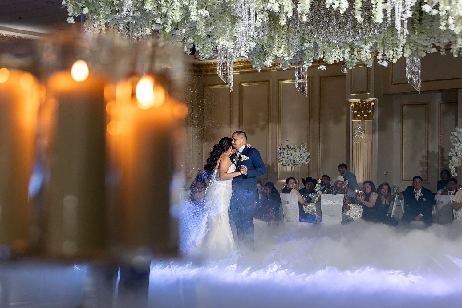 A bride and groom share their first dance on a cloud of low-lying fog under a ceiling of white flowers and crystal chandeliers.