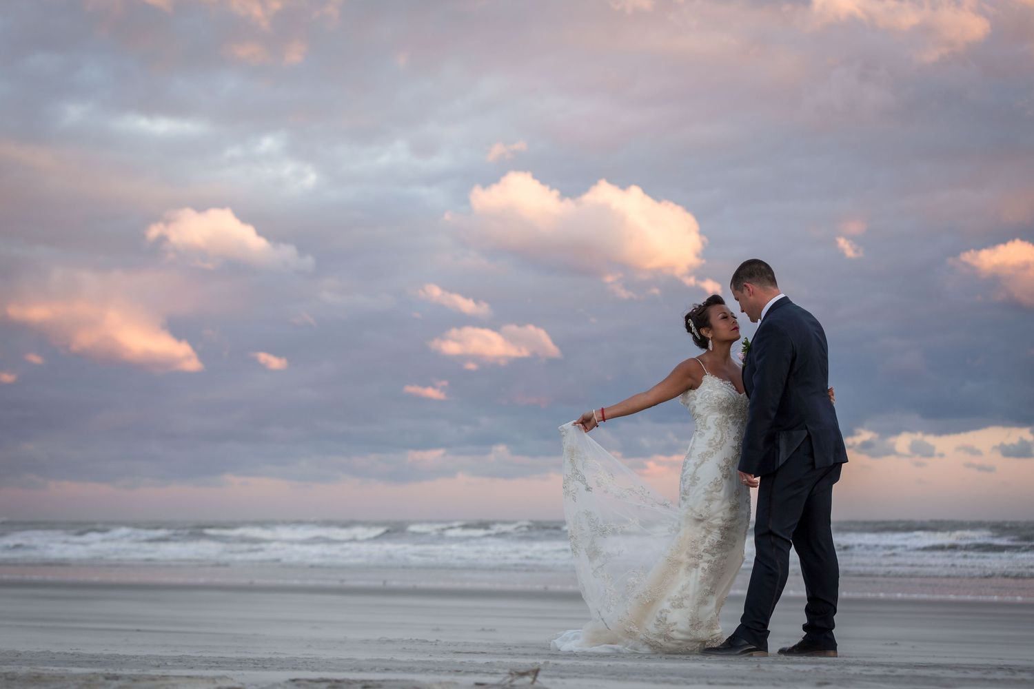 Bride and groom share a romantic moment on a beach at sunset with pink clouds and ocean waves in the background.
