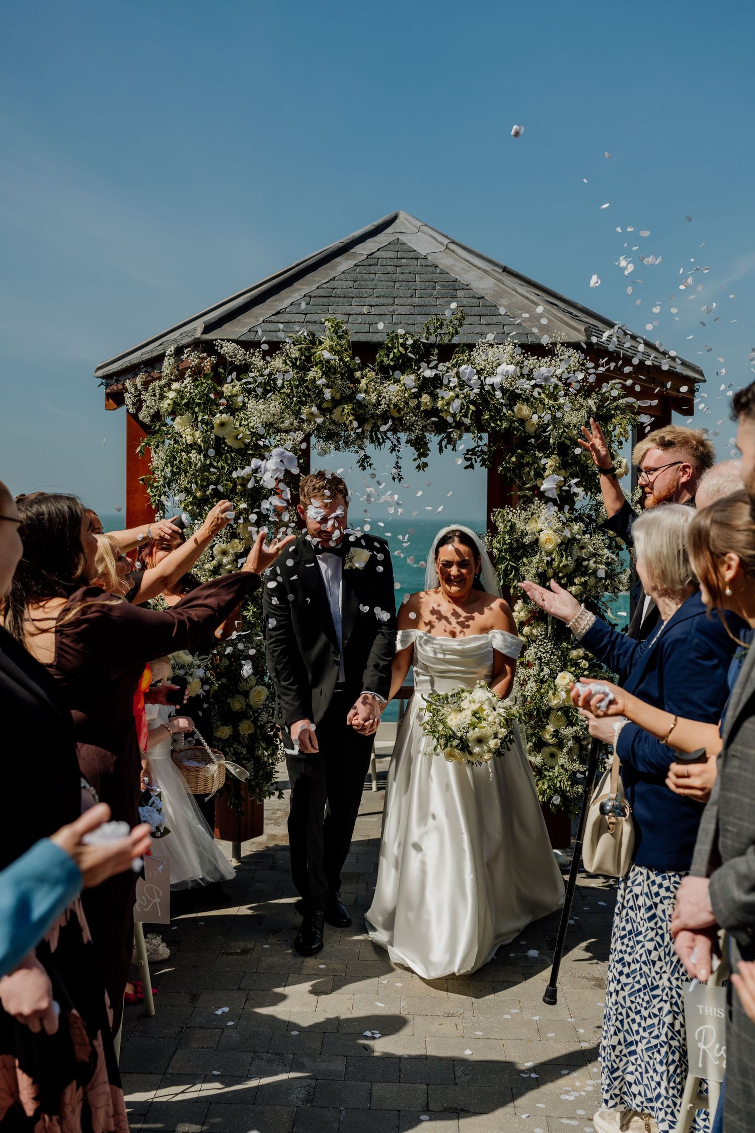 Bride and groom walk through confetti shower under floral arch gazebo on sunny wedding day.