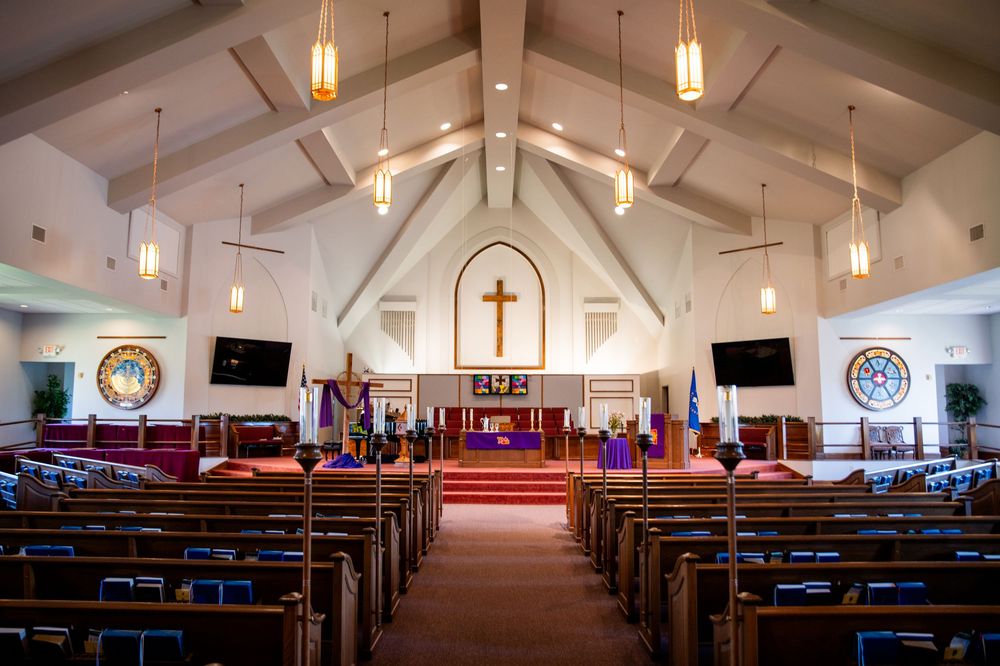 Bright church interior with wooden pews, vaulted ceiling, cross above altar, stained glass windows, and warm pendant lighting.